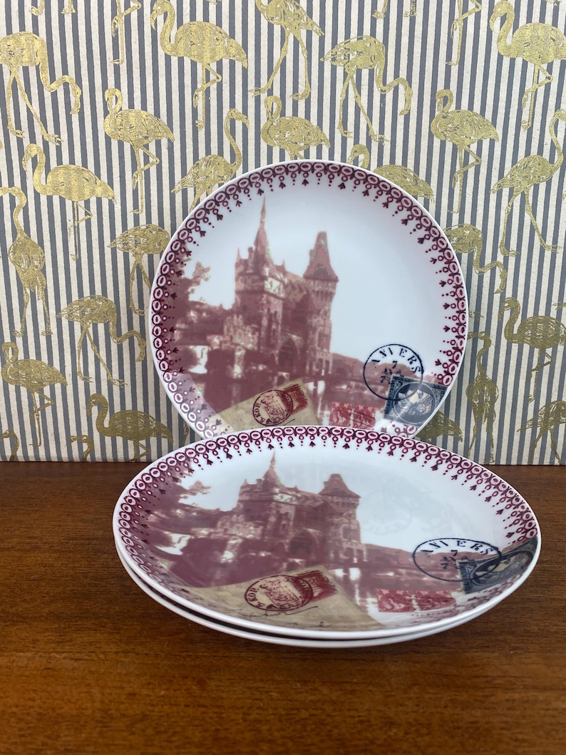 May include: Three white ceramic plates with a burgundy trim and a vintage image of a castle with a red roof and a brown background. The plates have a burgundy trim and a vintage image of a castle with a red roof and a brown background. The plates have a burgundy trim and a vintage image of a castle with a red roof and a brown background.