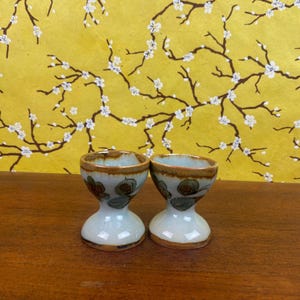 May include: Two ceramic egg cups with a light blue and brown glaze. Each cup features a flared base and a floral design in green and brown. The background is a yellow floral pattern.