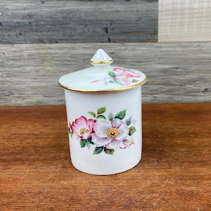 Pode incluir: Um pote de cerâmica branco com tampa, adornado com desenhos florais rosa e brancos e detalhes dourados. O pote é cilíndrico com uma forma ligeiramente curva. A tampa tem um pequeno pináculo pontiagudo e detalhes dourados. O padrão floral inclui folhas verdes.
