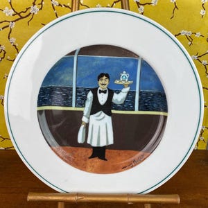 May include: A white ceramic plate with a green border features a painting of a waiter holding a tray with a teapot and cups. The waiter is dressed in a black vest and white apron. The background depicts a blue seascape.
