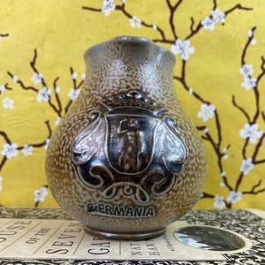 May include: A small, speckled ceramic pitcher with a decorative emblem. The emblem features a figure and the word "GERMANIA" in black lettering. The pitcher has a brown and tan color scheme and a spout for pouring.