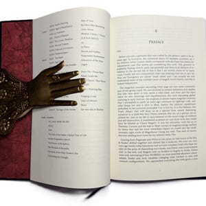May include: An open book with a decorative bronze hand bookmark. The bookmark is shaped like a hand with intricate details. The book has red and white pages with text. The book is open to the preface.