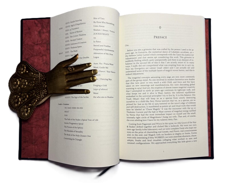 May include: An open book with a decorative bronze hand bookmark. The bookmark is shaped like a hand with intricate details. The book has red and white pages with text. The book is open to the preface.