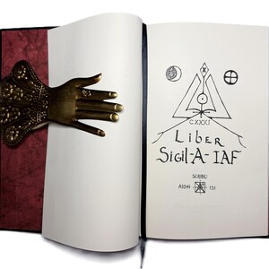 May include: An open book with a decorative bronze hand bookmark. The book's pages are white, with the title "Liber Sigil-A-IAF" and other text in black ink. The cover is black with a burgundy textured spine.