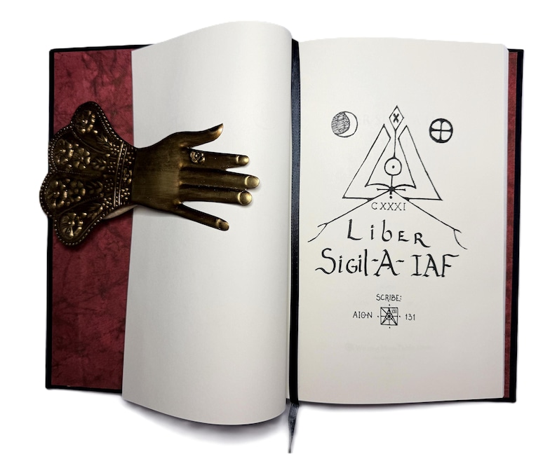 May include: An open book with a decorative bronze hand bookmark. The book's pages are white, with the title "Liber Sigil-A-IAF" and other text in black ink. The cover is black with a burgundy textured spine.