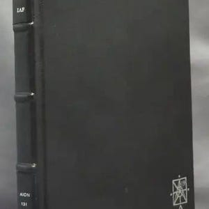 May include: A dark gray book with the title "Liber Sigil A IAP" and the number "131" printed on the spine. A white geometric symbol is on the lower right corner of the cover. The book has a textured appearance.