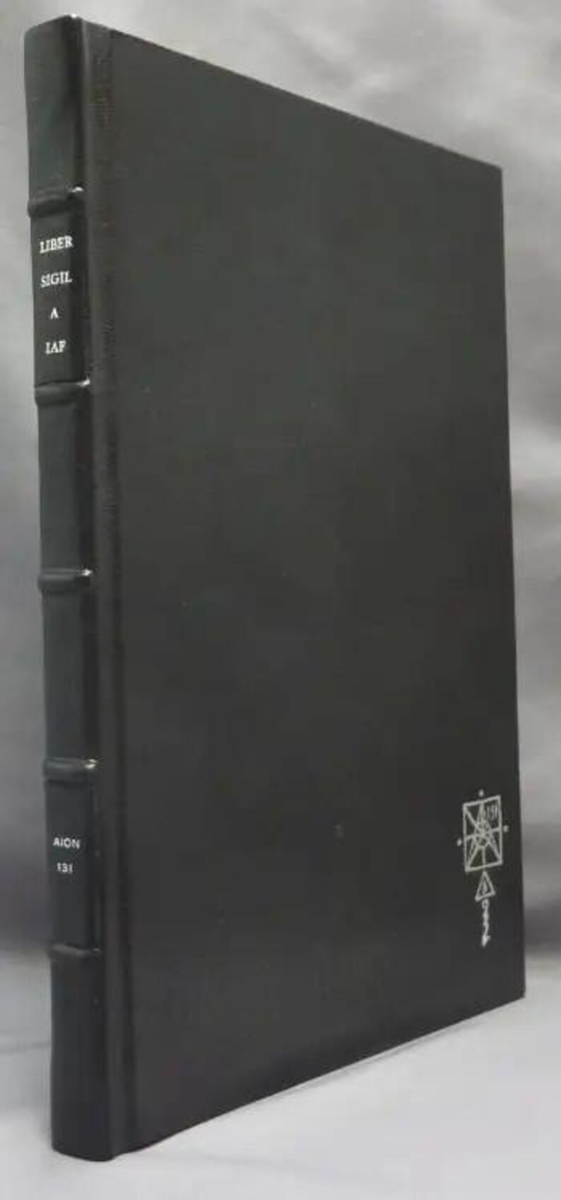 May include: A dark gray book with the title "Liber Sigil A IAP" and the number "131" printed on the spine. A white geometric symbol is on the lower right corner of the cover. The book has a textured appearance.