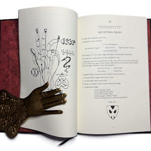 May include: An open book with illustrations and text, featuring a bronze hand-shaped bookmark. The book's pages display drawings of hands and symbols, with the title "ACCEPTING SIGILS" visible.