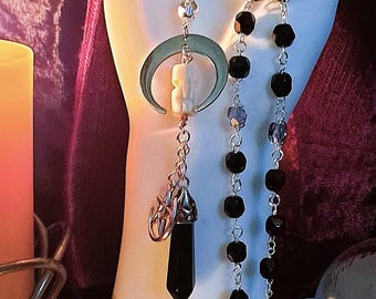May include: A silver chain necklace with a black crystal pendant, a crescent moon charm, and a bone charm. The necklace is on a white hand with black beads on a silver chain.