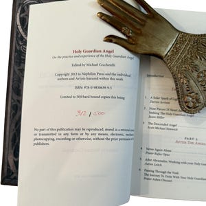 K&ouml;nnte beinhalten: Ein offenes Buch mit dem Titel "Holy Guardian Angel" mit einem dekorativen Buchseitenhalter in Form einer Messinghand. Der Titel und der Autor des Buches sind sichtbar, ebenso wie die ISBN und die limitierte Auflage 312/500.