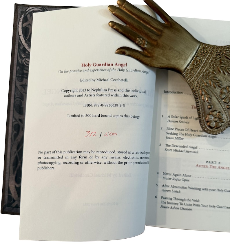 K&ouml;nnte beinhalten: Ein offenes Buch mit dem Titel "Holy Guardian Angel" mit einem dekorativen Buchseitenhalter in Form einer Messinghand. Der Titel und der Autor des Buches sind sichtbar, ebenso wie die ISBN und die limitierte Auflage 312/500.