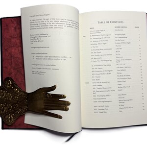 May include: An open book with a red cover and a decorative brass hand bookmark. The book's pages display text, including a table of contents. The bookmark is detailed with floral patterns and a ring on one finger.