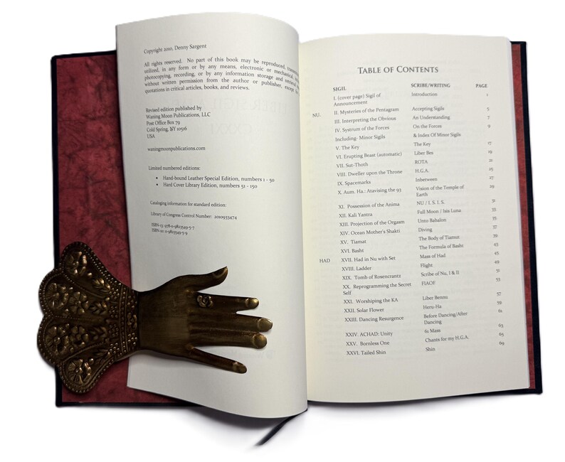 May include: An open book with a red cover and a decorative brass hand bookmark. The book's pages display text, including a table of contents. The bookmark is detailed with floral patterns and a ring on one finger.