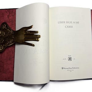 May include: An open book with the text "Liber Sigil A IAF" and a decorative brass hand bookmark. The bookmark is shaped like a hand with intricate details and rings on the fingers. The book has a red cover.