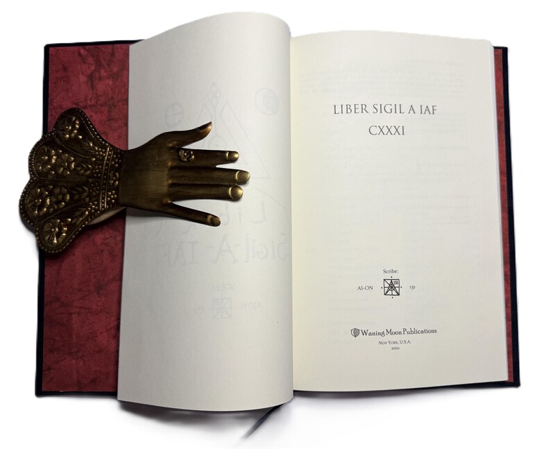May include: An open book with the text "Liber Sigil A IAF" and a decorative brass hand bookmark. The bookmark is shaped like a hand with intricate details and rings on the fingers. The book has a red cover.