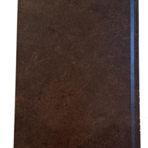 K&ouml;nnte beinhalten: Ein braunes Notizbuch mit strukturiertem Einband und einem blauen Streifen entlang des R&uuml;ckens. Ein cremefarbenes Lesezeichen ist unten sichtbar. Das Notizbuch ist wahrscheinlich zum Schreiben oder Notizen machen.