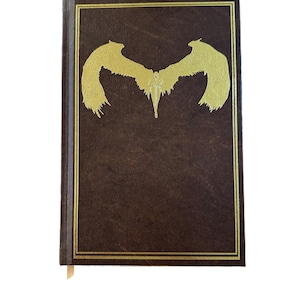 K&ouml;nnte beinhalten: Ein braunes Hardcover-Buch mit einem goldgepr&auml;gten Design eines stilisierten gefl&uuml;gelten Wesens. Der Einband hat einen goldenen Rand. Ein cremefarbenes Lesezeichen ist sichtbar.