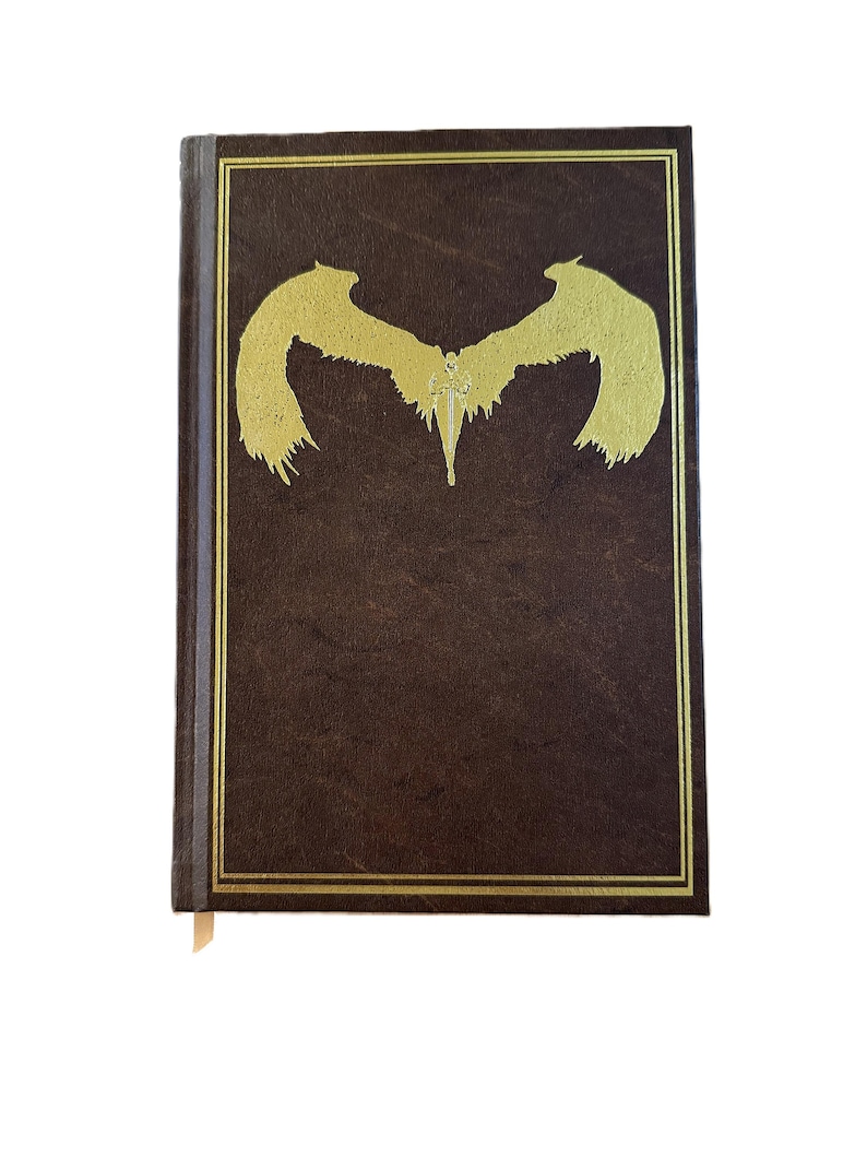 K&ouml;nnte beinhalten: Ein braunes Hardcover-Buch mit einem goldgepr&auml;gten Design eines stilisierten gefl&uuml;gelten Wesens. Der Einband hat einen goldenen Rand. Ein cremefarbenes Lesezeichen ist sichtbar.