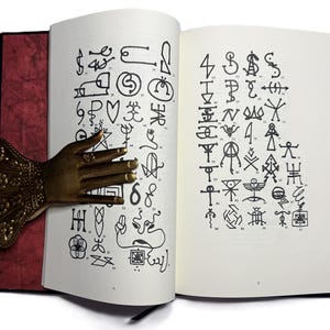 May include: An open book with pages displaying numerous black symbols and illustrations. A decorative bronze hand-shaped book holder is inserted between the pages. The book has a red cover and a black spine.