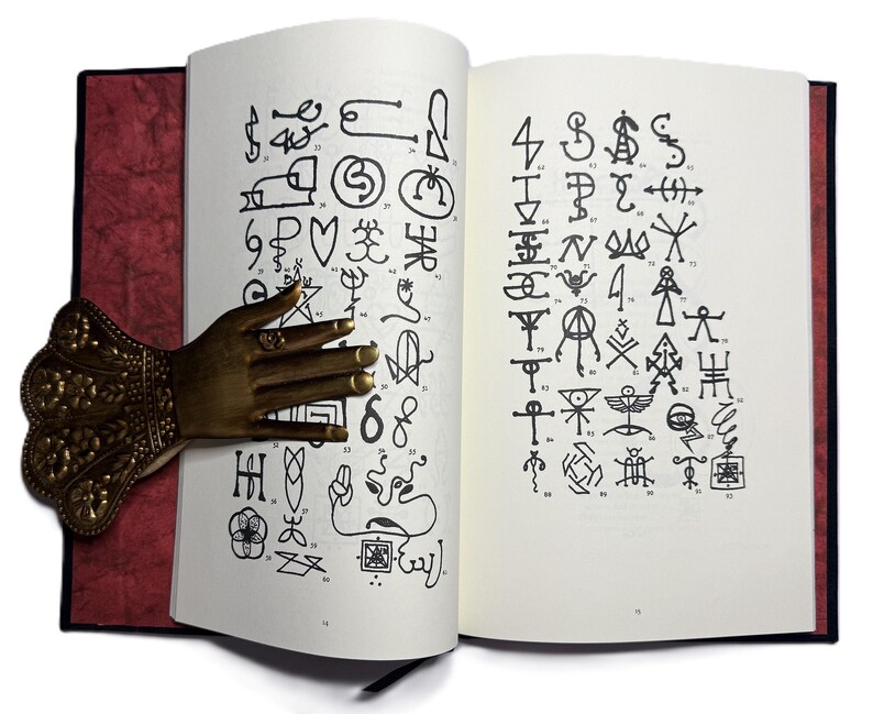 May include: An open book with pages displaying numerous black symbols and illustrations. A decorative bronze hand-shaped book holder is inserted between the pages. The book has a red cover and a black spine.