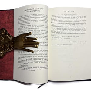 May include: An open book with text and a decorative brass hand bookmark. The bookmark is shaped like a hand with rings on the fingers. The book has red and black covers.