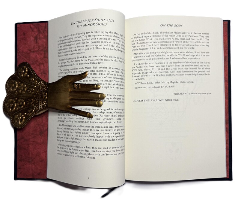 May include: An open book with text and a decorative brass hand bookmark. The bookmark is shaped like a hand with rings on the fingers. The book has red and black covers.