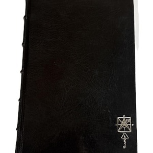 May include: A black leather-bound book with a textured cover. The spine is stitched with visible thread. A silver emblem and text are in the lower right corner. The book is closed and rectangular.