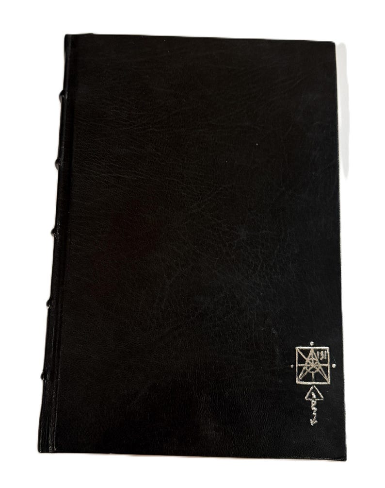 May include: A black leather-bound book with a textured cover. The spine is stitched with visible thread. A silver emblem and text are in the lower right corner. The book is closed and rectangular.