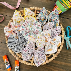 May include: A collection of fabric hair bows with school-themed patterns, including pencils, books, and letters, arranged in a woven basket. The bows are in various colors and patterns. A box of crayons, scissors, and glue sticks are also present.