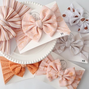 May include: Assortment of baby headbands in various colors and patterns. The headbands feature bows in shades of peach, orange, and white, with designs including stripes, dots, and suns. Some headbands have seashell and palm tree prints.