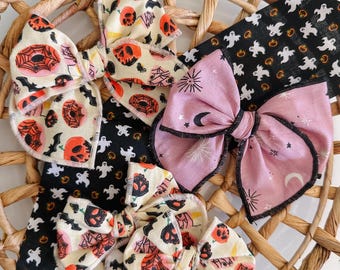 May include: Three fabric hair bows featuring Halloween patterns. One bow has a cream background with orange pumpkins, black bats, and spiderwebs. Another is light purple with black moons and stars. The third is a smaller cream bow with similar pumpkin motifs.