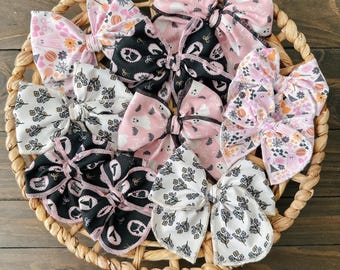 May include: A collection of fabric hair bows in a woven basket. The bows feature various Halloween-themed patterns, including ghosts, pumpkins, and floral designs, in colors like pink, black, white, and orange. The bows are arranged on a brown wooden surface.