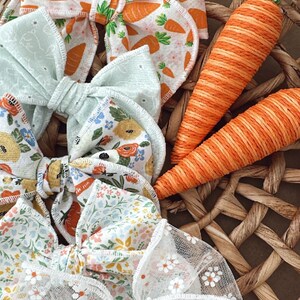 May include: A collection of colorful hair bows with floral and carrot patterns, alongside two orange carrot decorations, all arranged in a woven basket. The bows feature various designs, including white lace and pastel fabrics.