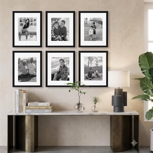 May include: A gallery wall arrangement of six black rectangular picture frames with white matting, each containing a black and white photograph. The frames are displayed in two rows of three, mounted on a wall above a long console table.