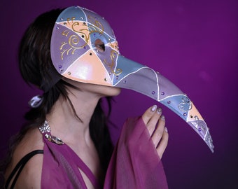 May include: A plague doctor mask featuring a long, pointed beak and a geometric pattern of lavender, peach, and slate blue segments. The mask is decorated with gold swirling line accents and small, embedded purple rhinestones along the seams.