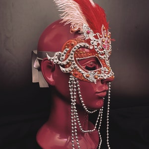 May include: A red and white feathered masquerade mask with silver and rhinestone embellishments. The mask is attached to a silver beaded chain.