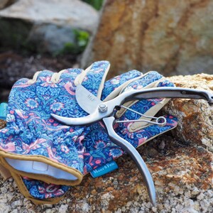 May include: A pair of blue gardening gloves with a floral pattern and a silver pruning shears with a black handle. The shears are labeled "Miel" and "Moor&Pen".