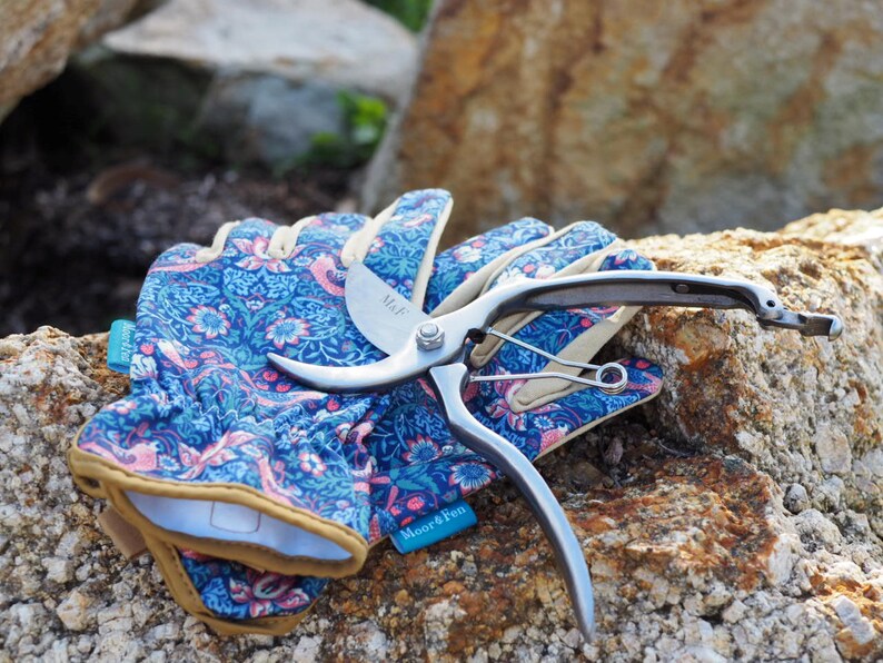 May include: A pair of blue gardening gloves with a floral pattern and a silver pruning shears with a black handle. The shears are labeled "Miel" and "Moor&Pen".