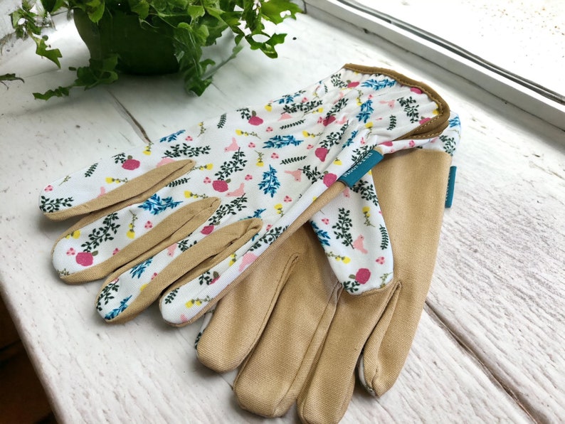 May include: A pair of gardening gloves with a floral print. The gloves are white with pink, blue, and yellow flowers and leaves. The palms of the gloves are a light brown color.
