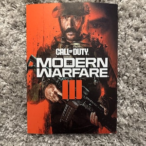 May include: A Call of Duty: Modern Warfare III poster featuring a soldier in a black tactical vest and helmet holding a rifle. The background is a bright orange colour with black splatters.