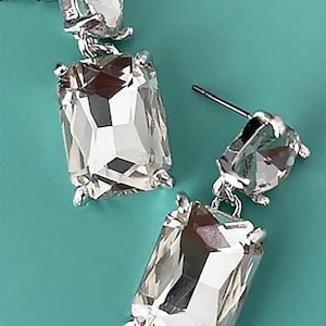 May include: A pair of silver-toned, dangling earrings with large, clear, rectangular-cut crystals. Each earring features a smaller, round crystal at the top and a larger, rectangular crystal below. The crystals reflect light, creating a sparkling effect.