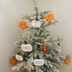 May include: A white artificial Christmas tree decorated with fall-themed ornaments. The ornaments are shaped like pumpkins and leaves and have various autumn-inspired words written on them, such as "Blessings", "Happy Fall", "Thankfull", "Cozy", "Football", "Snuggling", "Bonfire", "Carmel Apple", "Gather", "Harvest", "Pumpkin Spice", and "Eat More Pie".