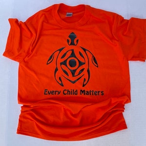 May include: Orange t-shirt with a black turtle design and the text "Every Child Matters". The shirt is made of a soft material and has short sleeves and a crew neck.
