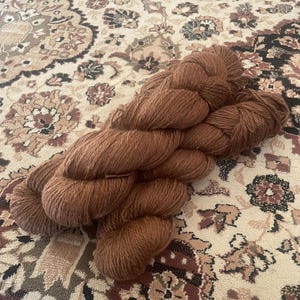 May include: A skein of brown yarn, likely wool or a similar fiber, rests on a patterned rug. The yarn is a warm, earthy tone, and the rug features a complex design with beige, brown, and burgundy elements. The yarn appears soft and ready for crafting.