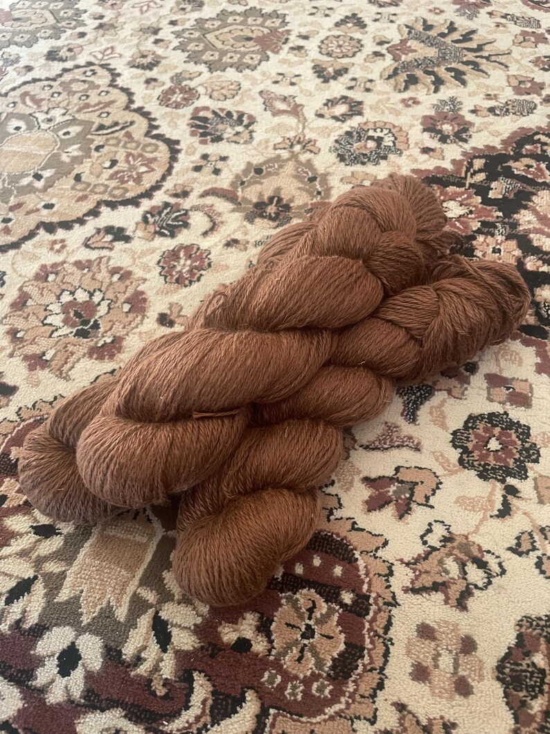 May include: A skein of brown yarn, likely wool or a similar fiber, rests on a patterned rug. The yarn is a warm, earthy tone, and the rug features a complex design with beige, brown, and burgundy elements. The yarn appears soft and ready for crafting.
