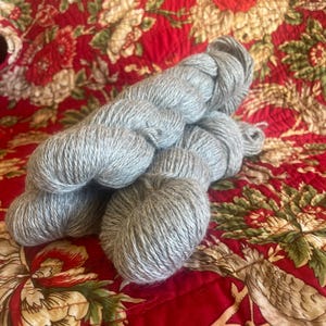 May include: Several skeins of light gray yarn are arranged on a red floral patterned fabric. The yarn appears soft and fluffy, ideal for knitting or crocheting projects. The fabric has a quilted texture.