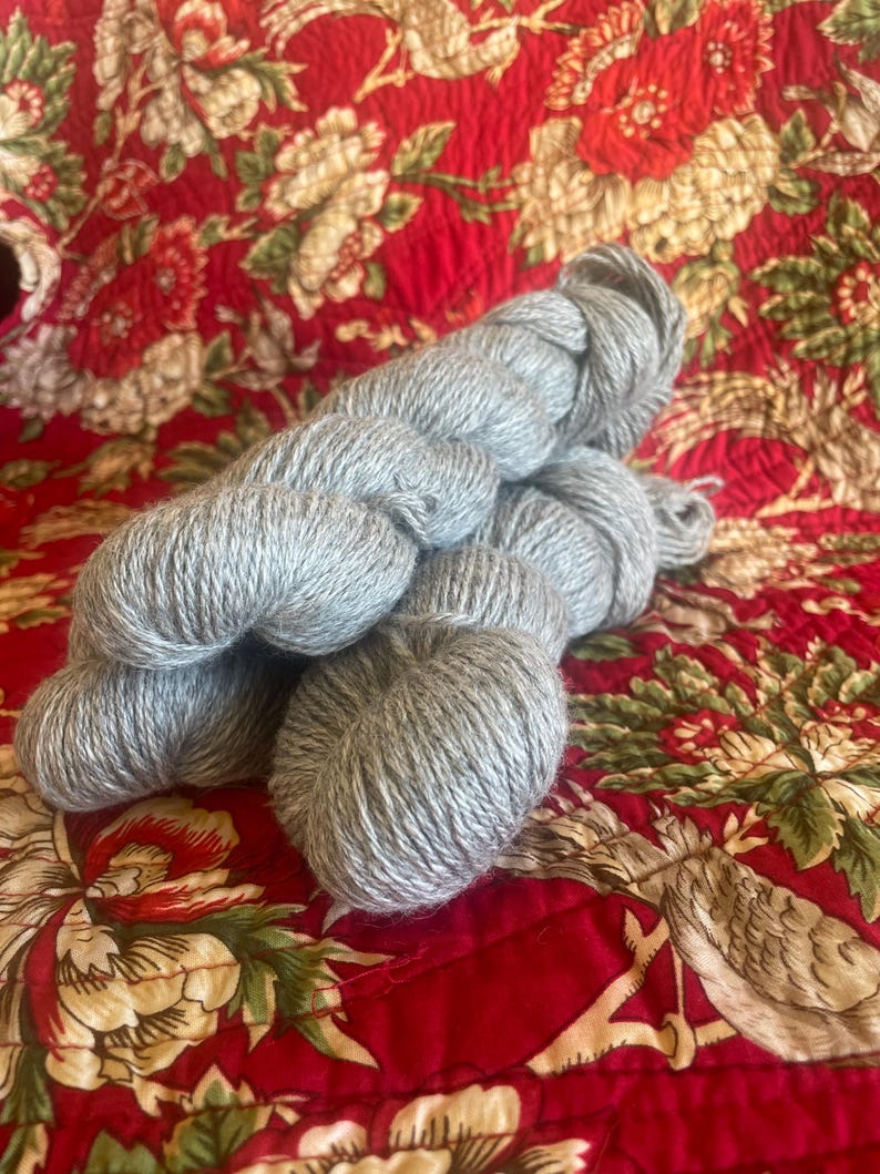 May include: Several skeins of light gray yarn are arranged on a red floral patterned fabric. The yarn appears soft and fluffy, ideal for knitting or crocheting projects. The fabric has a quilted texture.