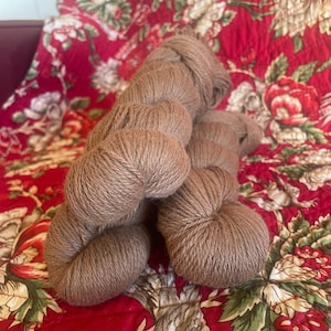May include: Several skeins of brown yarn are arranged on a red floral patterned fabric. The yarn appears to be soft and loosely spun, with a natural, earthy tone. The fabric has a vintage, quilted appearance.