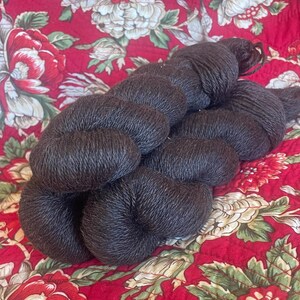 May include: Dark brown yarn skeins, tightly wound, resting on a red floral patterned fabric. The yarn appears soft and textured, ideal for knitting or crocheting projects. The fabric features large red and cream-colored flowers.