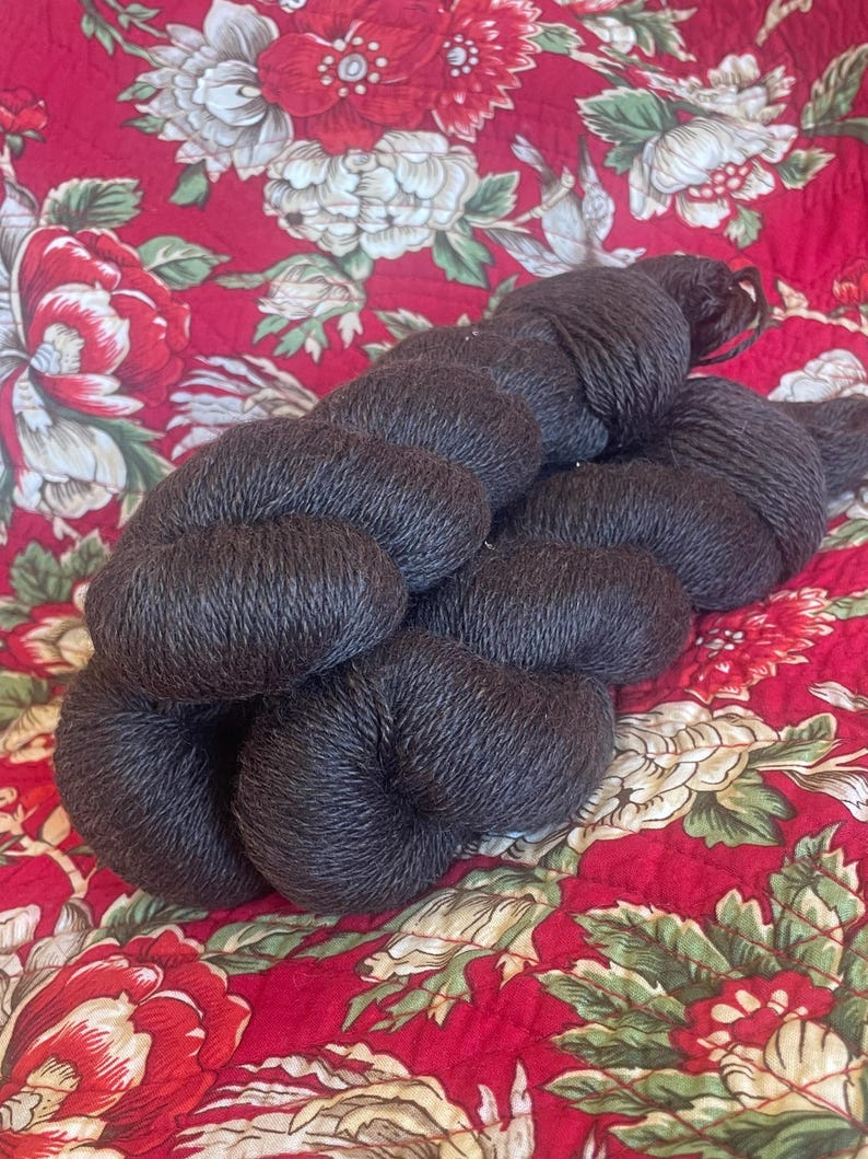 May include: Dark brown yarn skeins, tightly wound, resting on a red floral patterned fabric. The yarn appears soft and textured, ideal for knitting or crocheting projects. The fabric features large red and cream-colored flowers.